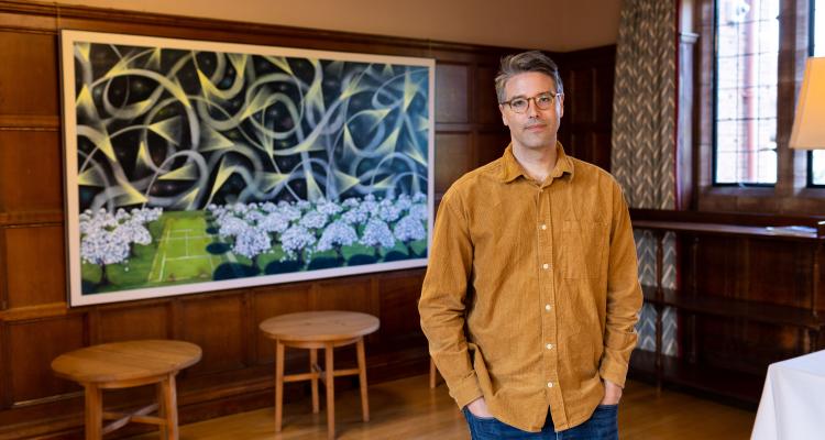 Luke Burton stands in front of his painting Impossible Weather, 2020. Oil and acrylic on canvas, 150 x 250 cm. Painting depicts the orchard and tennis court with stylised starry night sky above.