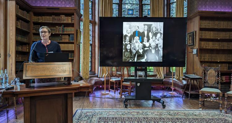 Prof Emma Gilby at a podium beside a screen with a black and white image of women